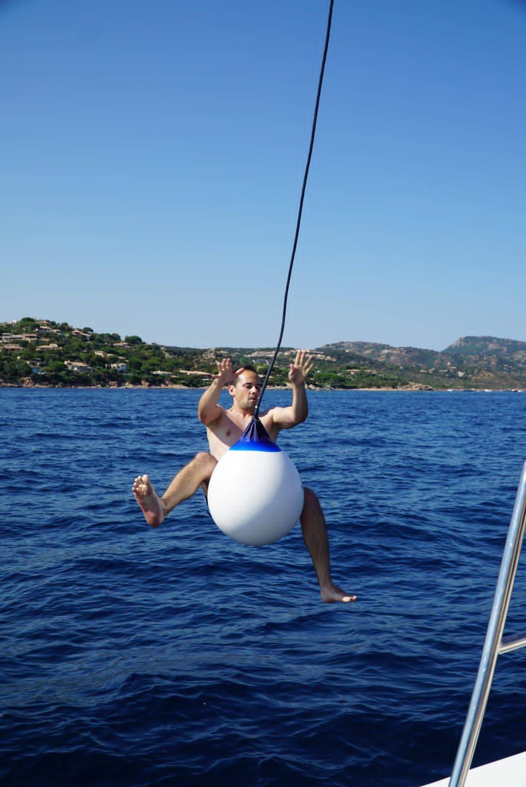 Ein Mann schaukelt während eines Segelurlaubs fröhlich auf einer großen weißen Boje, die an einer Yacht hängt, mit dem blauen Meer und der hügeligen Küste im Hintergrund.