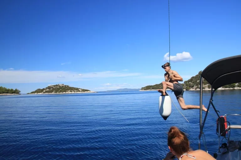 Ein Mann fährt während eines Segelurlaubs mit dem Wakeboard von einem Boot ins klare blaue Wasser unter einem sonnigen Himmel, mit entfernten Inseln im Hintergrund.