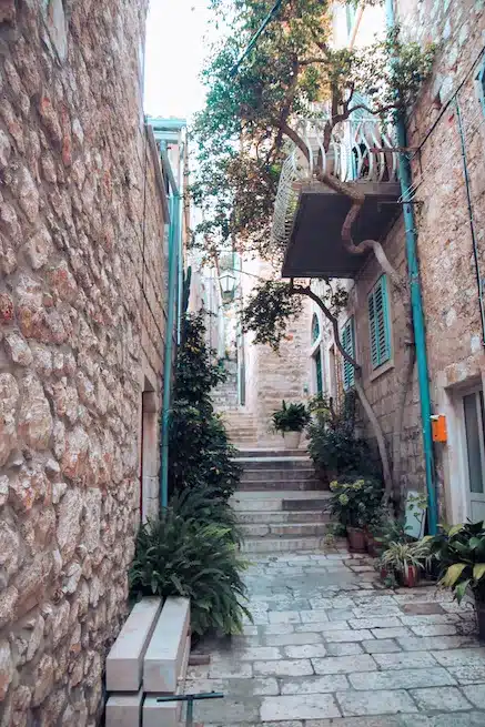 Schmale Gasse mit Steinmauern, Kopfsteinpflasterstufen und üppigen grünen Pflanzen in einer malerischen europäischen Altstadtkulisse, perfekt für ein Segeln-Abenteuer.