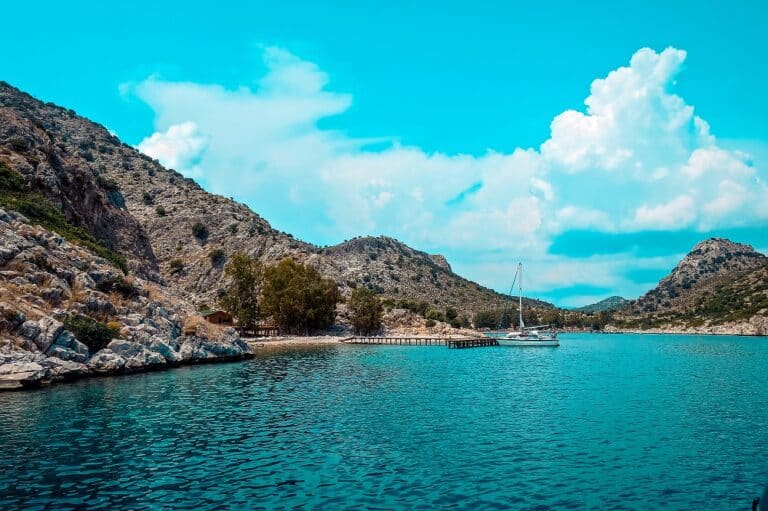 Eine ruhige Küstenszene mit einer Segelyacht, die auf ruhigem blauem Wasser in der Nähe einer felsigen Küste mit grünen Hügeln unter einem wolkenverhangenen Himmel schwimmt. Ein kleiner Holzsteg erstreckt sich vom Ufer ins Meer