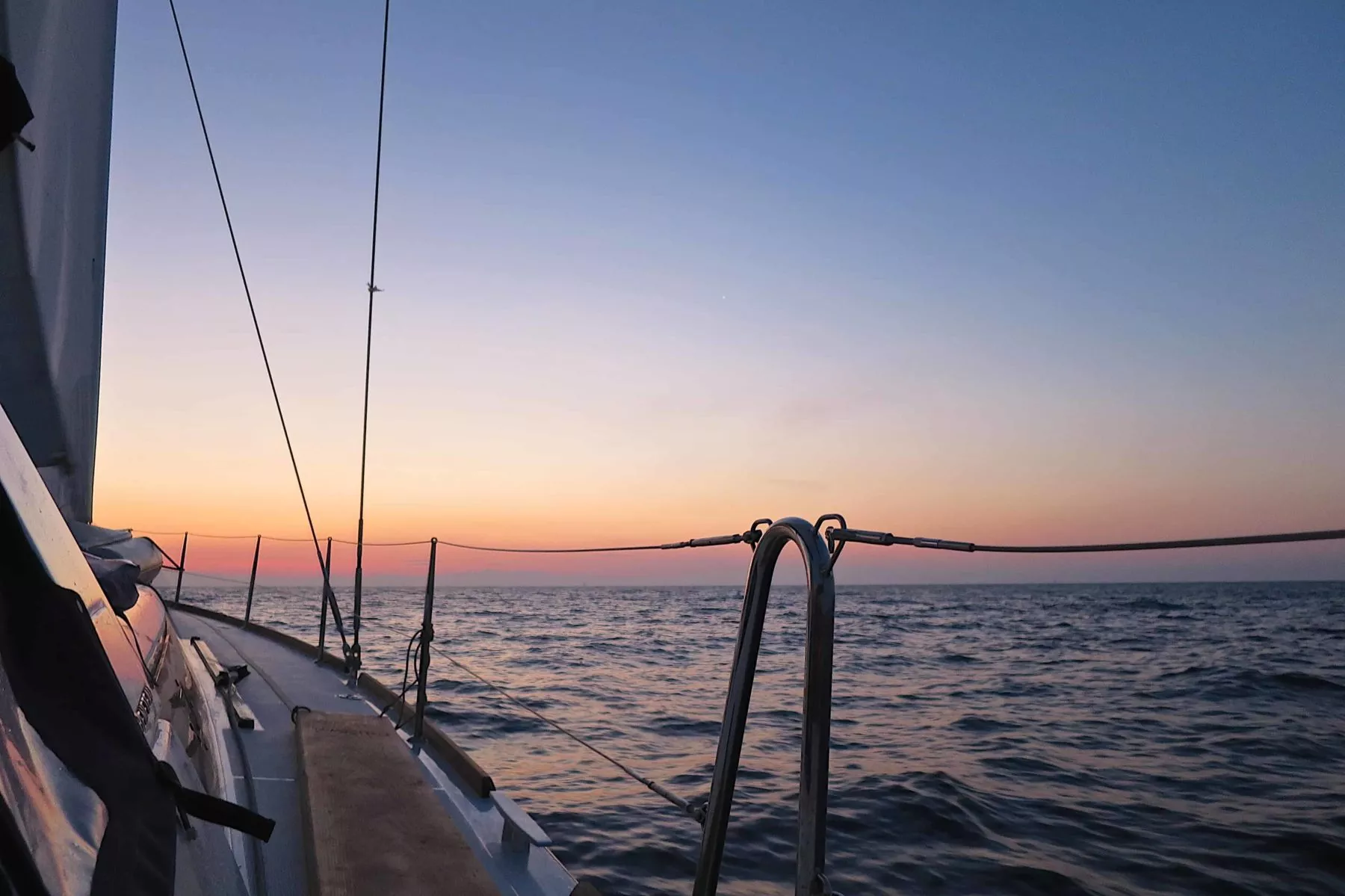 Sonnenuntergang vom Deck eines Segelboots während eines Segeltörns aus gesehen. Zu sehen sind die Reling des Bootes und ein Teil des Segels vor dem Hintergrund einer ruhigen See und eines farbenprächtigen Himmels.