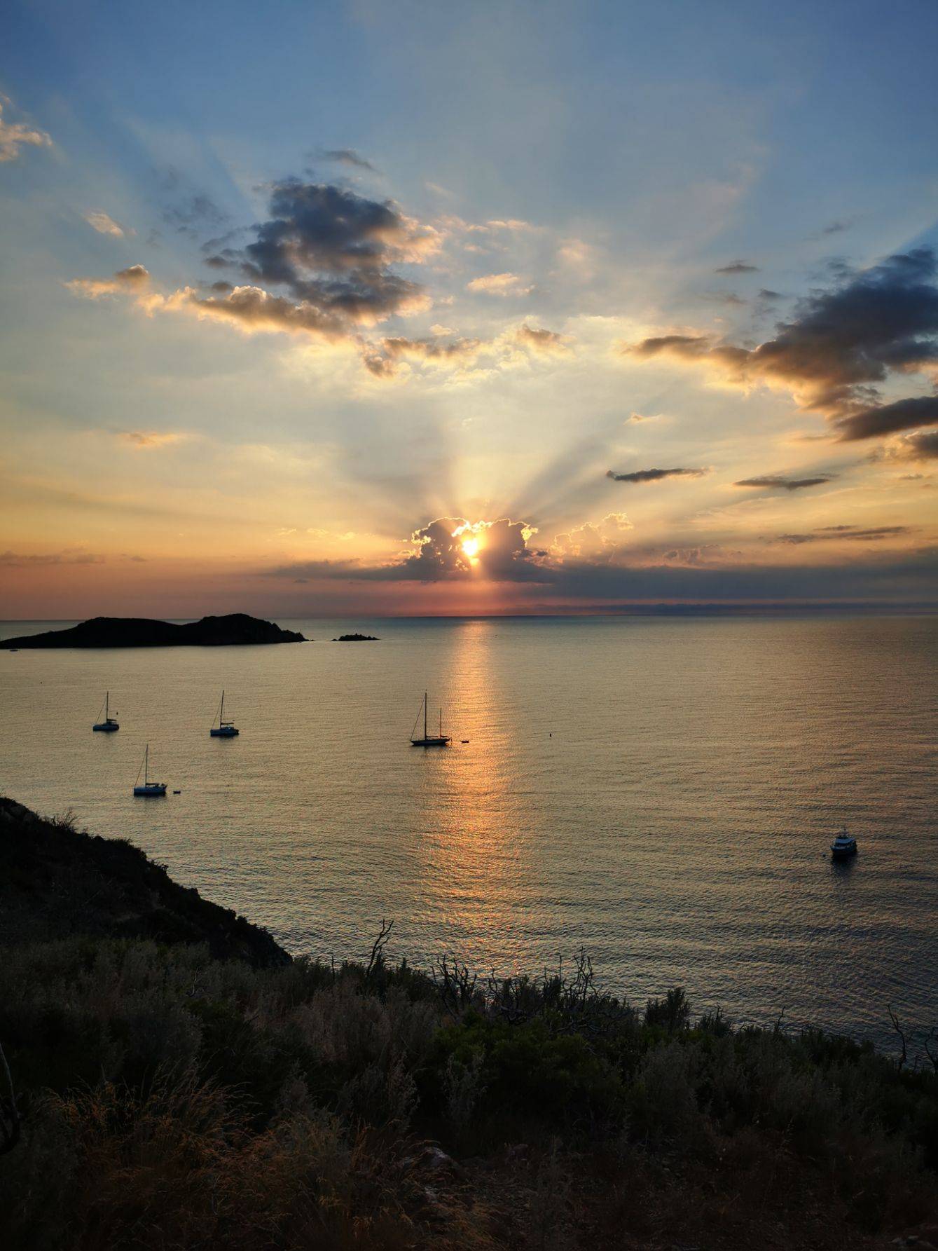Sonnenuntergang über dem Meer mit Sonnenstrahlen, die durch die Wolken brechen und auf das Wasser scheinen. Mehrere Segelyachten liegen in Küstennähe vor Anker, eingerahmt von der Landschaftssilhouette.