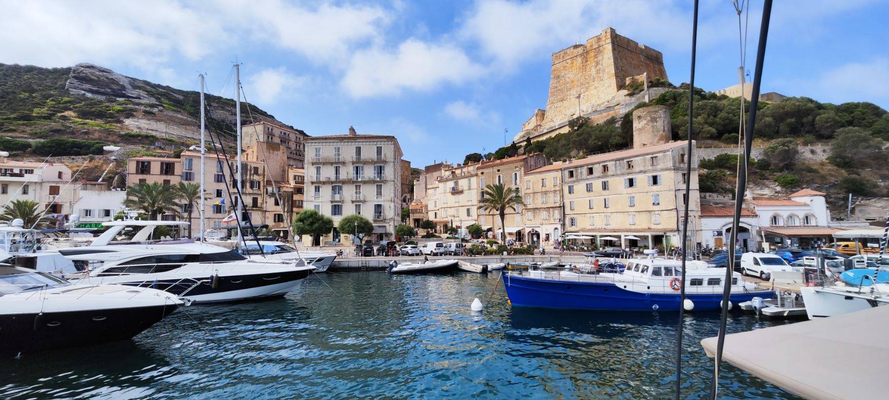 Luxusyachten, die in einem ruhigen Hafen mit mehrstöckigen pastellfarbenen Gebäuden und einer großen historischen Festung auf einem Hügel unter einem klaren blauen Himmel vor Anker liegen, perfekt für einen Segelurlaub.