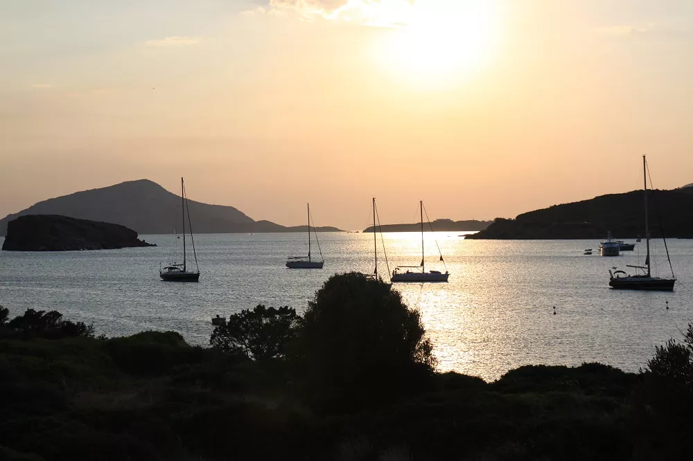 Ein malerischer Sonnenuntergang über einer ruhigen Bucht mit mehreren Segelbooten, die in der Nähe einer grünen, hügeligen Küste vor Anker liegen und während ihres Segelurlaubs Silhouetten gegen die helle Sonne bilden, die sich im Wasser spiegelt.