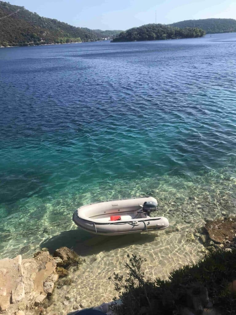 Eine kleine Segelyacht, die im klaren, türkisfarbenen Wasser in der Nähe einer felsigen Küste vor Anker liegt, mit einem üppigen grünen Wald im Hintergrund unter einem klaren, blauen Himmel.