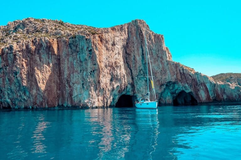Ein weißes Segelboot ankert in der Nähe einer großen Felsklippe mit mehreren Meereshöhlen unter einem klaren blauen Himmel. Das Wasser ist ruhig und azurblau und spiegelt die sonnenbeschienenen Klippen während eines ruhigen Segelurlaubs wider