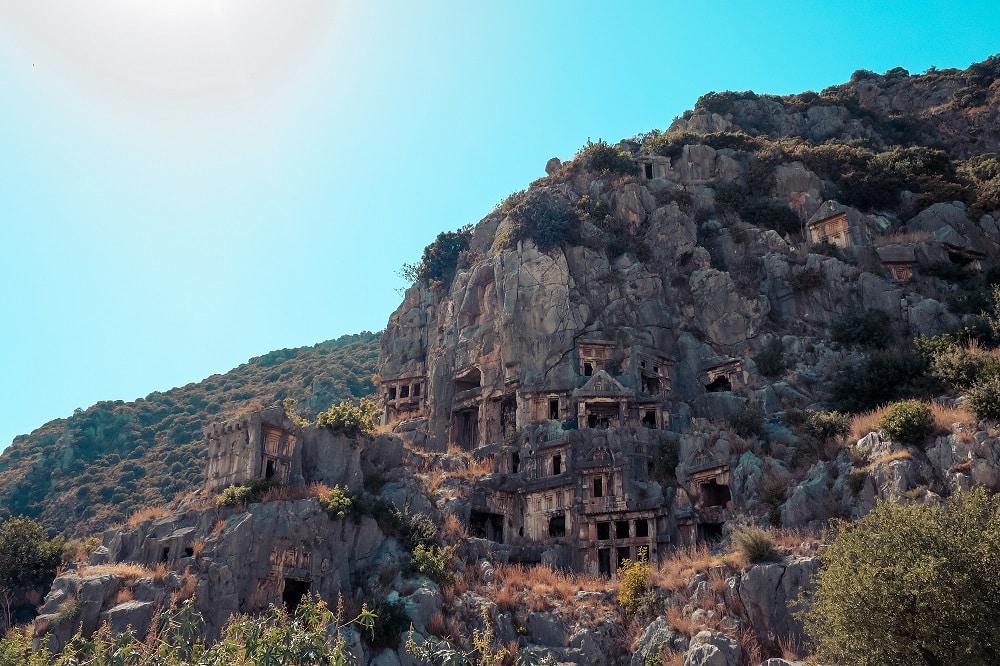 Uralte, in den Felsen gehauene Gräber, eingebettet in einen schroffen Hügel unter einem klaren blauen Himmel, zeigen eindrucksvolle historische Architektur in einer natürlichen Bergkulisse, sichtbar von einem ruhigen Segeltörn.
