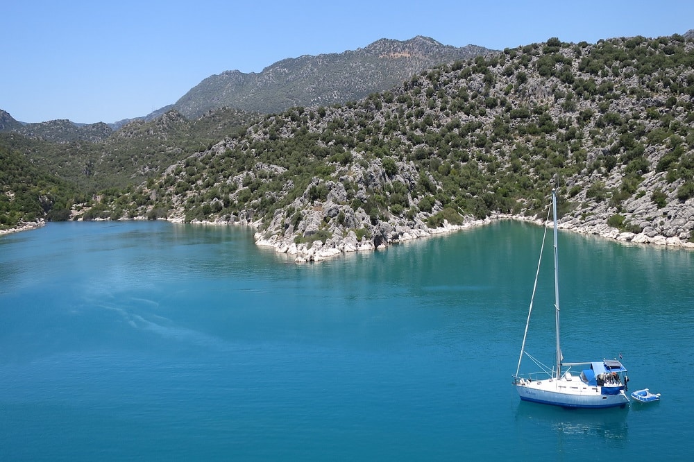 Eine ruhige, türkisfarbene Bucht, umgeben von felsigen Hügeln mit grünen Gipfeln. Ein einsames weißes Segelboot liegt im klaren Wasser vor Anker und unterstreicht die ruhige und malerische Naturlandschaft, die perfekt für einen Segeltörn ist.