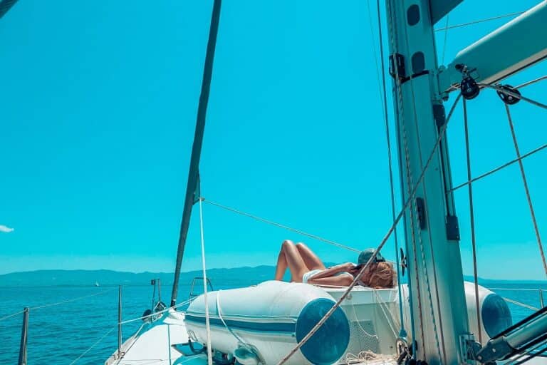 Eine Person entspannt sich auf dem Deck eines Segelboots, genießt die Sonne, über ihr den blauen Himmel und rundherum die Weite des Ozeans, während das Boot auf einem klaren Segeltörn dahinsegelt.