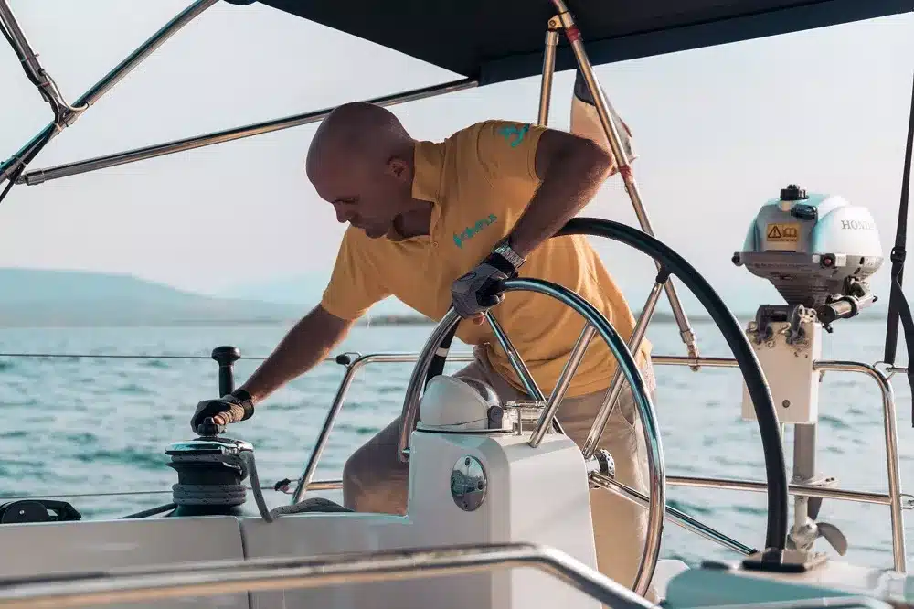 Ein Mann steuert eine Segelyacht und stellt die Winde ein, während er an einem klaren Tag am Steuer steht, mit dem Ozean im Hintergrund.