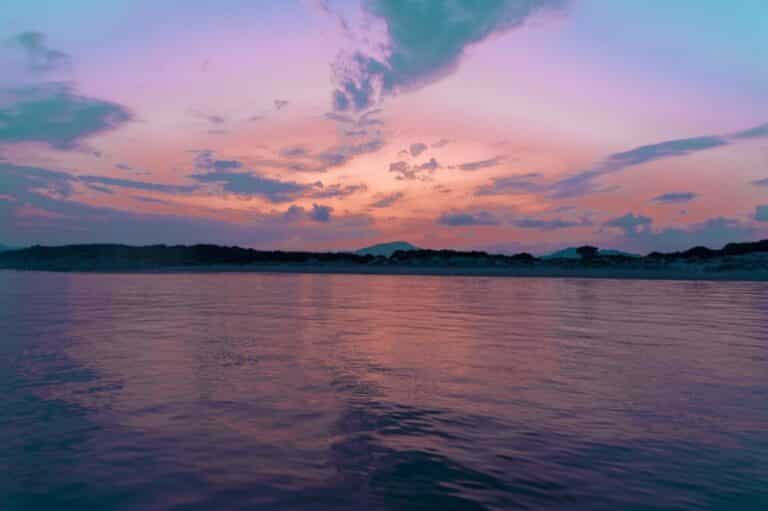 Eine ruhige Szene eines ruhigen Meeres unter einem lebendigen Sonnenuntergang mit Schattierungen von Rosa und Lila am Himmel und einer Spiegelung auf dem Wasser, mit einer Segelyacht-Silhouette in der Ferne