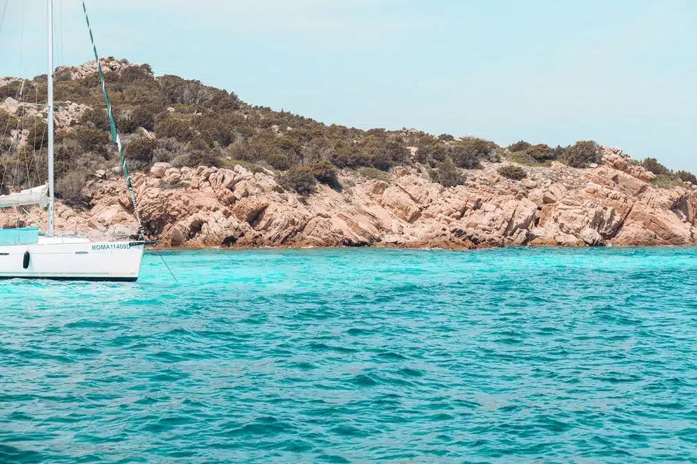 Eine ruhige Küstenszene mit einem weißen Segelboot, das auf leuchtend türkisfarbenem Wasser in der Nähe einer felsigen Küste mit üppigem Grün unter einem klaren Himmel segelt.