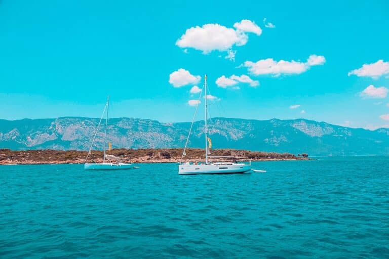 Zwei Segelboote ankern während ihres Segelurlaubs unter einem klaren blauen Himmel in der Nähe einer felsigen Küste, im Hintergrund sind in der Ferne Berge zu sehen.