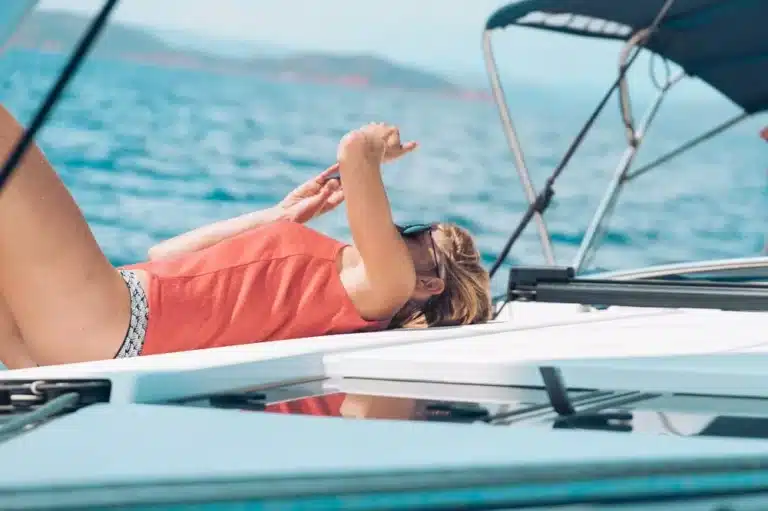 Eine Frau entspannt sich auf dem Deck eines Segelboots, sonnt sich und schützt ihre Augen mit der Hand, umgeben vom offenen Wasser.