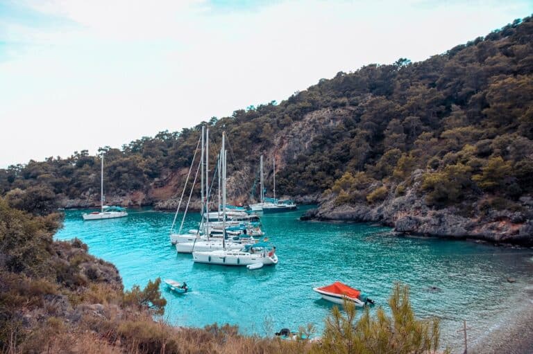Malerischer Blick auf eine ruhige Bucht mit kristallklarem, türkisfarbenem Wasser, umgeben von üppigen grünen Hügeln. Mehrere Segelboote und kleinere Boote, darunter eine Segelyacht, liegen im ruhigen Wasser vor Anker.