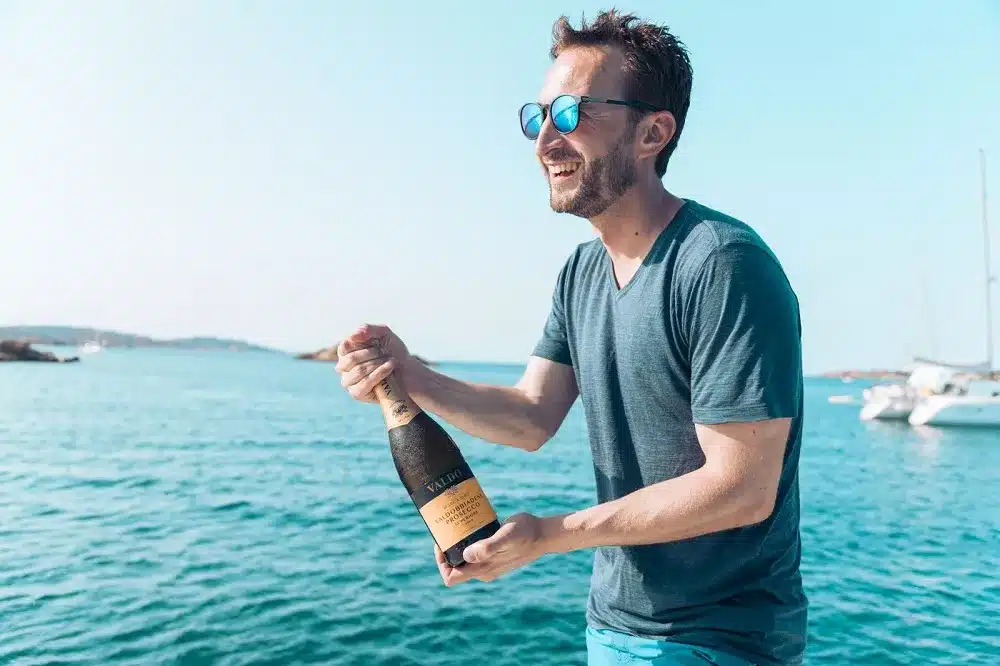 Ein fröhlicher Mann hält eine Champagnerflasche am Meer, mit Segelyachten im Hintergrund an einem sonnigen Tag. Er trägt eine blaue Sonnenbrille und ein graues T-Shirt und lächelt breit.