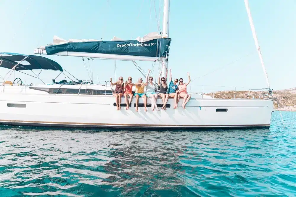 Sieben Personen sitzen auf der Seite einer Segelyacht und winken fröhlich in die Kamera. Die Yacht liegt auf ruhigem, blauem Wasser und man sieht einen klaren Himmel. Der Name des Bootes, Traum