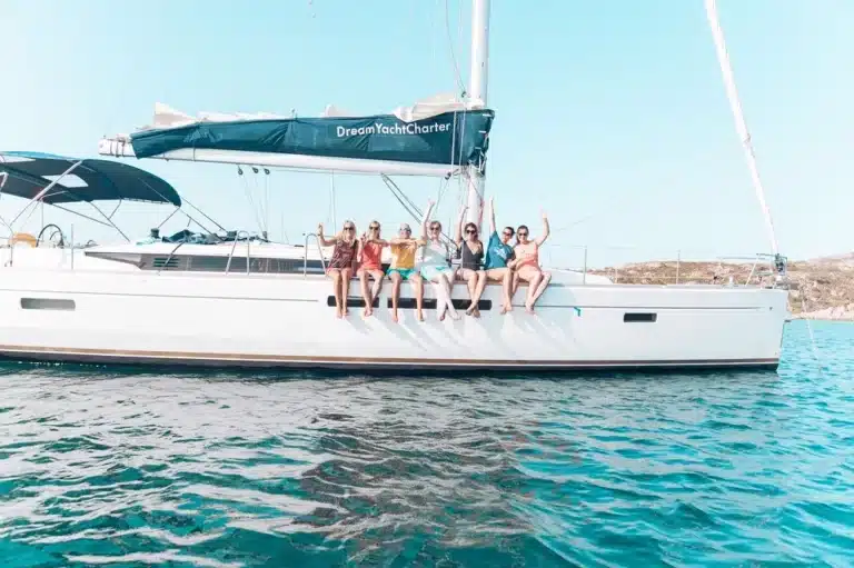 Eine Gruppe von sechs Freunden sitzt an der Seite einer weißen Segelyacht und jubelt, während die Yacht während ihres Segeltörns unter einem sonnigen Himmel im klaren blauen Wasser vor Anker liegt.