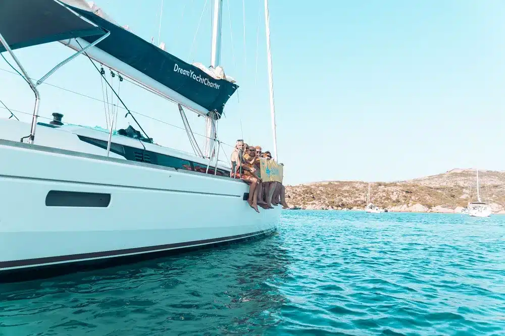 Eine Gruppe von Menschen mit Schwimmwesten steht an der Seite einer weißen Segelyacht, die unter einem strahlenden Himmel im klaren, türkisfarbenen Wasser in der Nähe einer felsigen Küste vor Anker liegt.