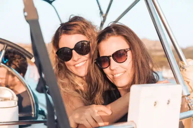 Zwei Frauen mit Sonnenbrillen lächeln und machen an einem sonnigen Tag ein Selfie auf einem Segeltörn mit offenem Verdeck. Auf ihren Gesichtern ist deutlich Freude und Kameradschaft zu erkennen.