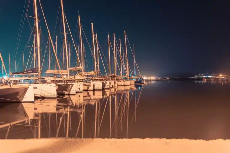 Nachtansicht eines ruhigen Yachthafens mit mehreren nebeneinander angedockten Segelyachten, die sich unter einem Sternenhimmel im Wasser spiegeln.