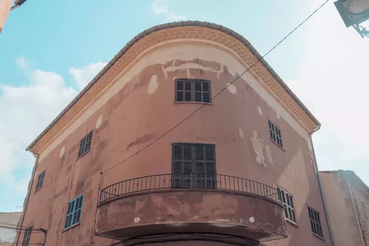 Ein verwittertes pfirsichfarbenes Gebäude mit abgerundeter Eckfassade, hölzernen Fensterläden und einem kleinen Balkon vor einem klaren blauen Himmel, der an die Aussicht während eines Segelurlaubs erinnert.