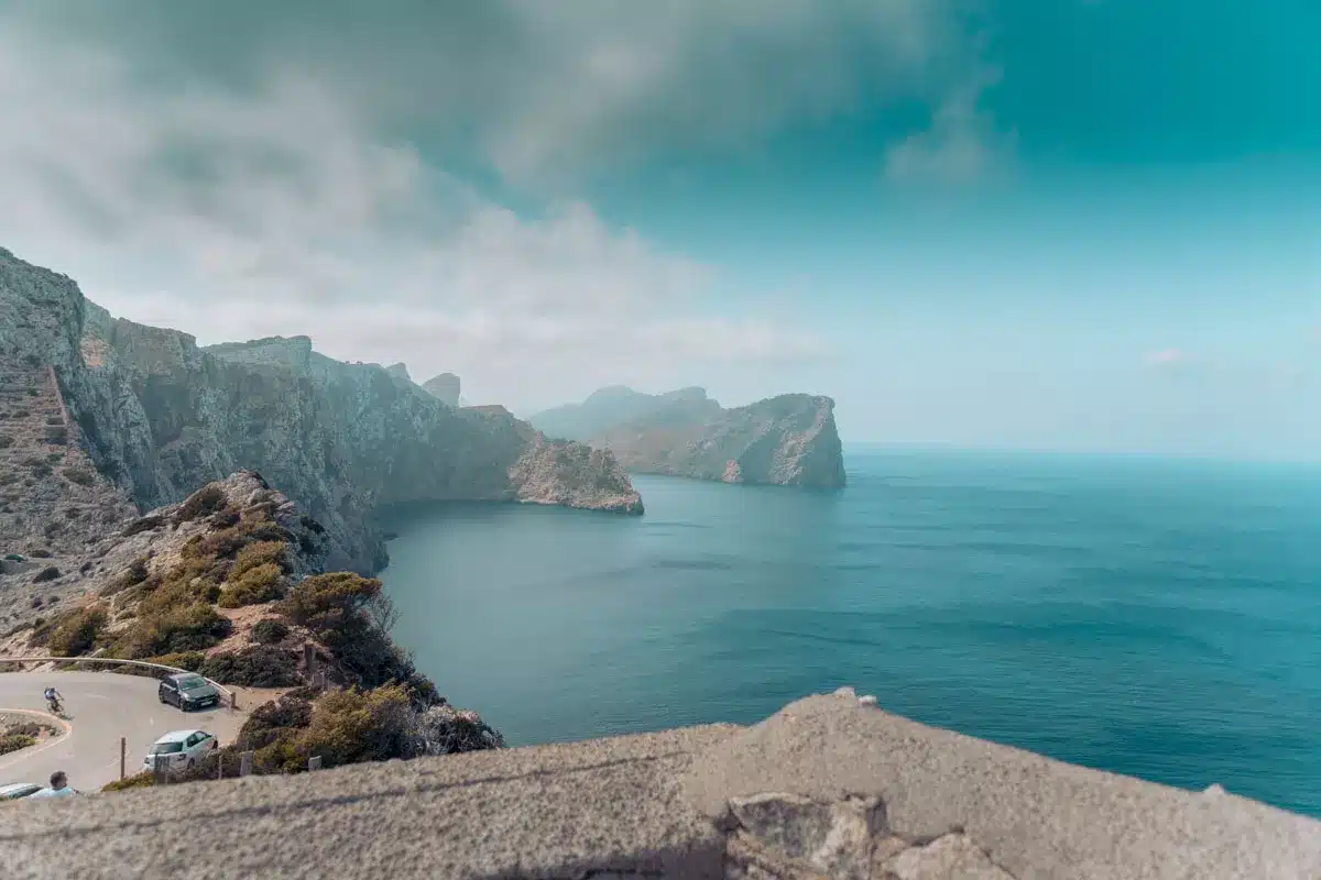 Eine malerische Küstenansicht mit einer kurvenreichen Straße entlang einer schroffen Klippe mit tiefblauem Meer auf der einen Seite und felsigen Bergen unter einem teilweise bewölkten Himmel, perfekt für die Planung eines Segelurlaubs.