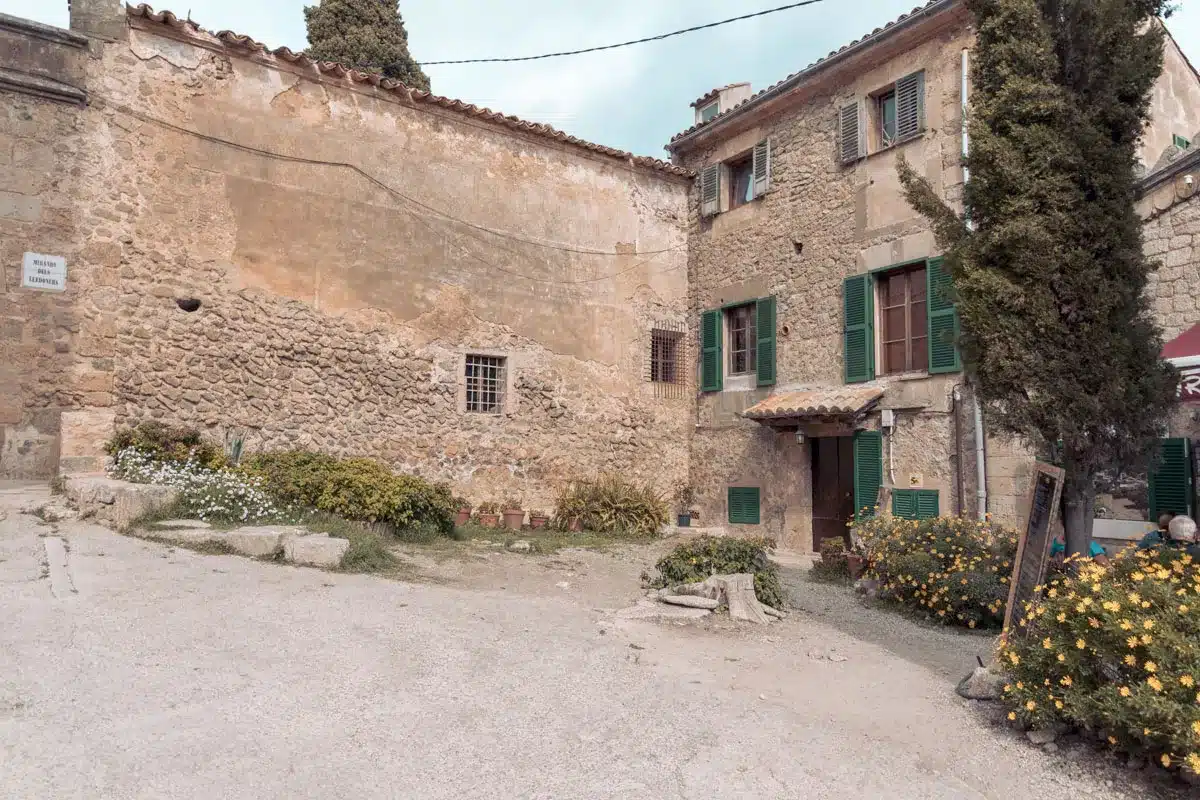 Eine malerische, rustikale Dorfecke mit alten Steinhäusern, grünen Fensterläden und einer gepflasterten Straße, die mit blühenden Sträuchern geschmückt ist. Eine ruhige und malerische Altstadtszene, perfekt für einen entspannten Segeltörn.