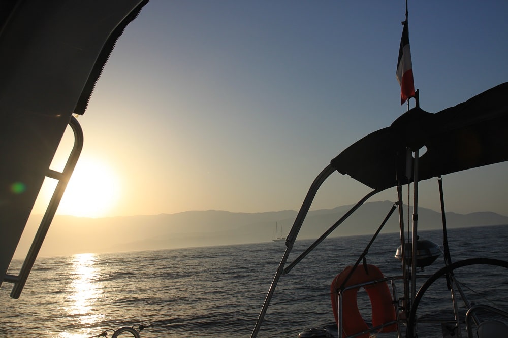 Blick von einer Segelyacht bei Sonnenuntergang. Zu sehen ist die Silhouette des Bootshecks mit Rettungsring und Flagge auf ein ruhiges Meer mit Bergen in der Ferne und einem weiteren Segelboot.
