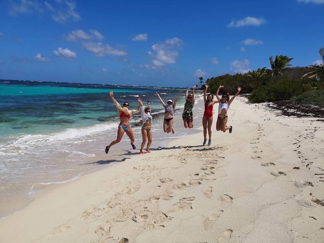 Sechs Menschen springen fröhlich an einem Sandstrand herum, im Hintergrund das Meer und eine Segelyacht unter einem klaren blauen Himmel.