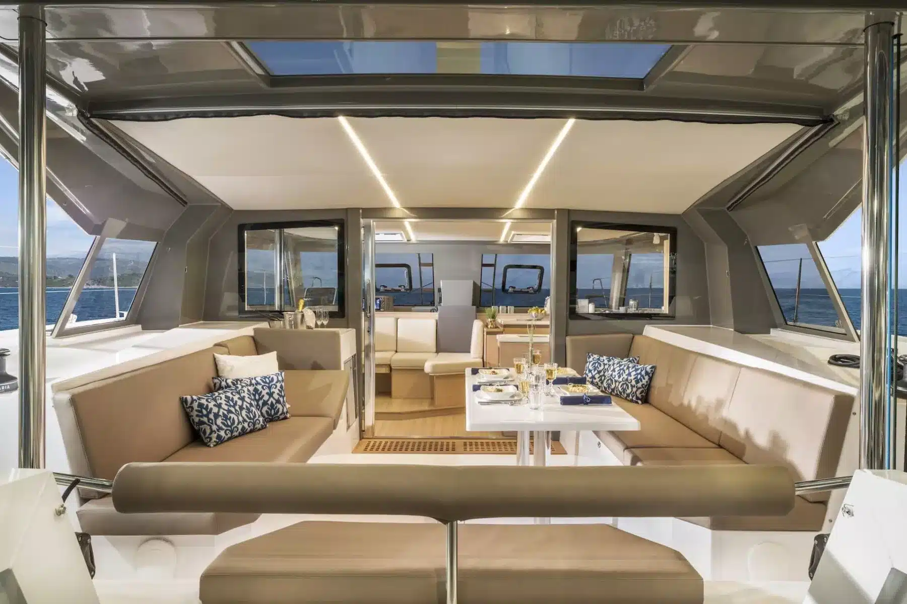 Innenraum einer luxuriösen Segelyacht mit bequemen beigen Sitzgelegenheiten, einem gedeckten Esstisch und großen Fenstern, die für reichlich Tageslicht sorgen.