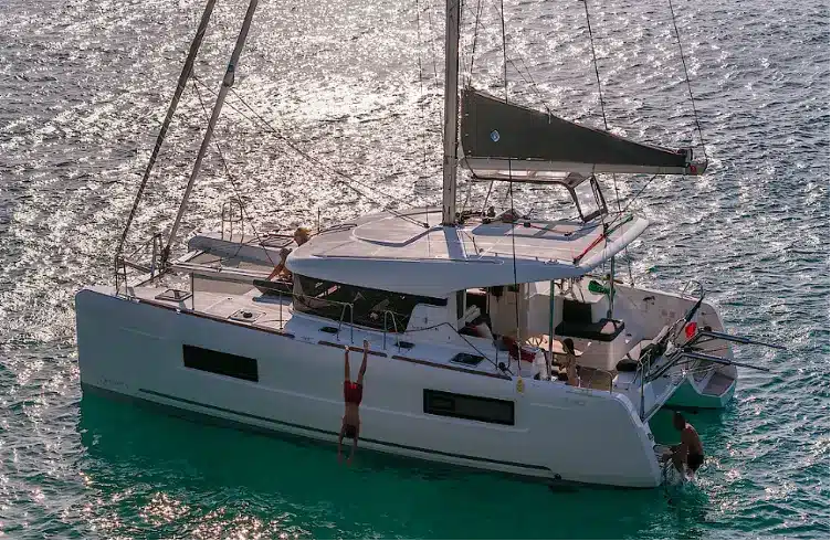 Ein weißer Katamaran mit zwei Personen, die einen sonnigen Tag auf dem Meer genießen; eine Person steht auf dem Achterdeck, während die andere während ihrer Segelre unter der Markise des Bootes sitzt