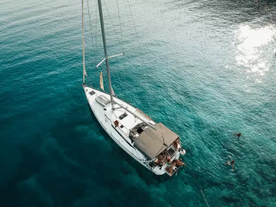 Luftaufnahme einer weißen Segelyacht auf einem klaren, blauen Meer mit Menschen, die auf dem Deck ein Sonnenbad nehmen, und einer Person, die während ihres Segeltörns in der Nähe schwimmt. Badeurlaub auf dem Segelboot: Die schönsten Buchten entdecken