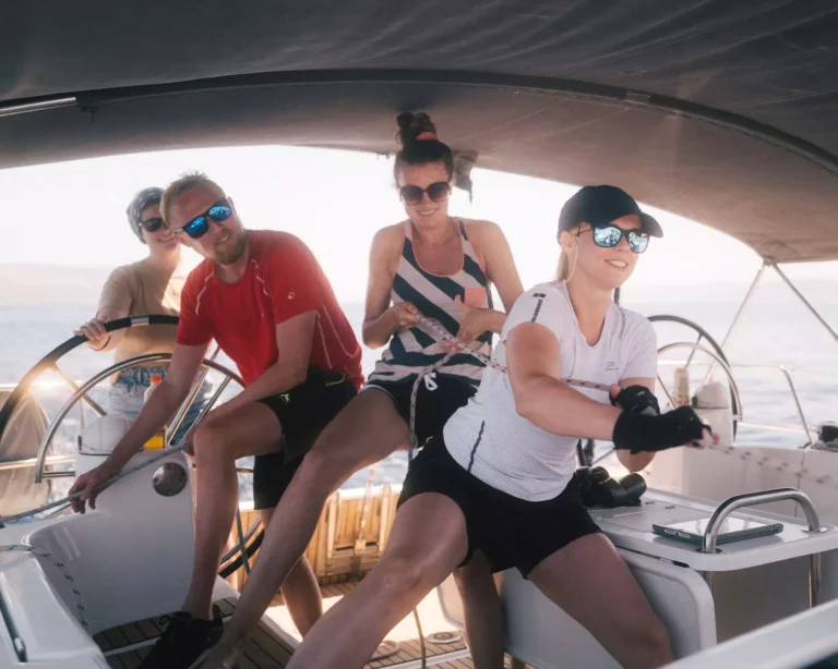 Vier Freunde genießen einen Segeltörn; sie tragen lässige Sommeroutfits, Sonnenbrillen und Hüte und zeigen Teamwork und entspannte Lächeln an einem sonnigen Tag auf dem Meer.