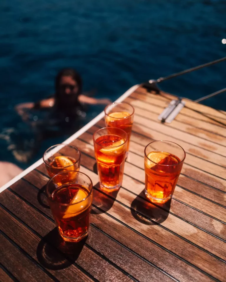 Vier Gläser Eistee mit Zitronenscheiben auf dem Holzdeck einer Yacht, mit Blick auf eine Person, die während einer Segelreise im klaren, blauen Ozeanwasser schwimmt.