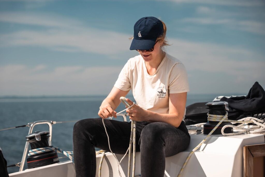 Eine junge Frau mit Mütze und Sonnenbrille sitzt auf einem Boot und lächelt, während sie während ihres Segelurlaubs einen Knoten in ein Seil bindet, im Hintergrund der klare Himmel und das ruhige Meer.