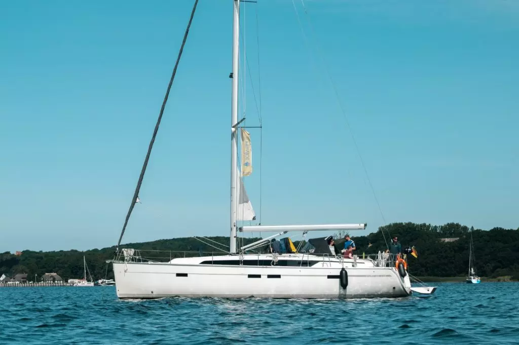 Eine weiße Segelyacht mit einer Gruppe von Menschen an Bord schwimmt auf ruhigem blauen Wasser unter einem klaren Himmel. – Bild.
