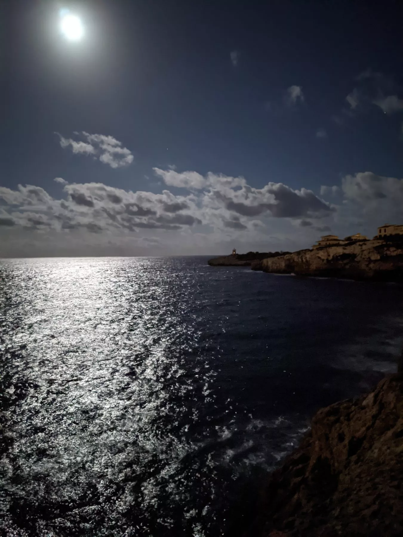 Mondbeschienene Meereslandschaft mit einem leuchtenden Mond, der sich im Ozean spiegelt, mit schroffen Klippen auf der rechten Seite unter einem Sternenhimmel während einer Segeltour.