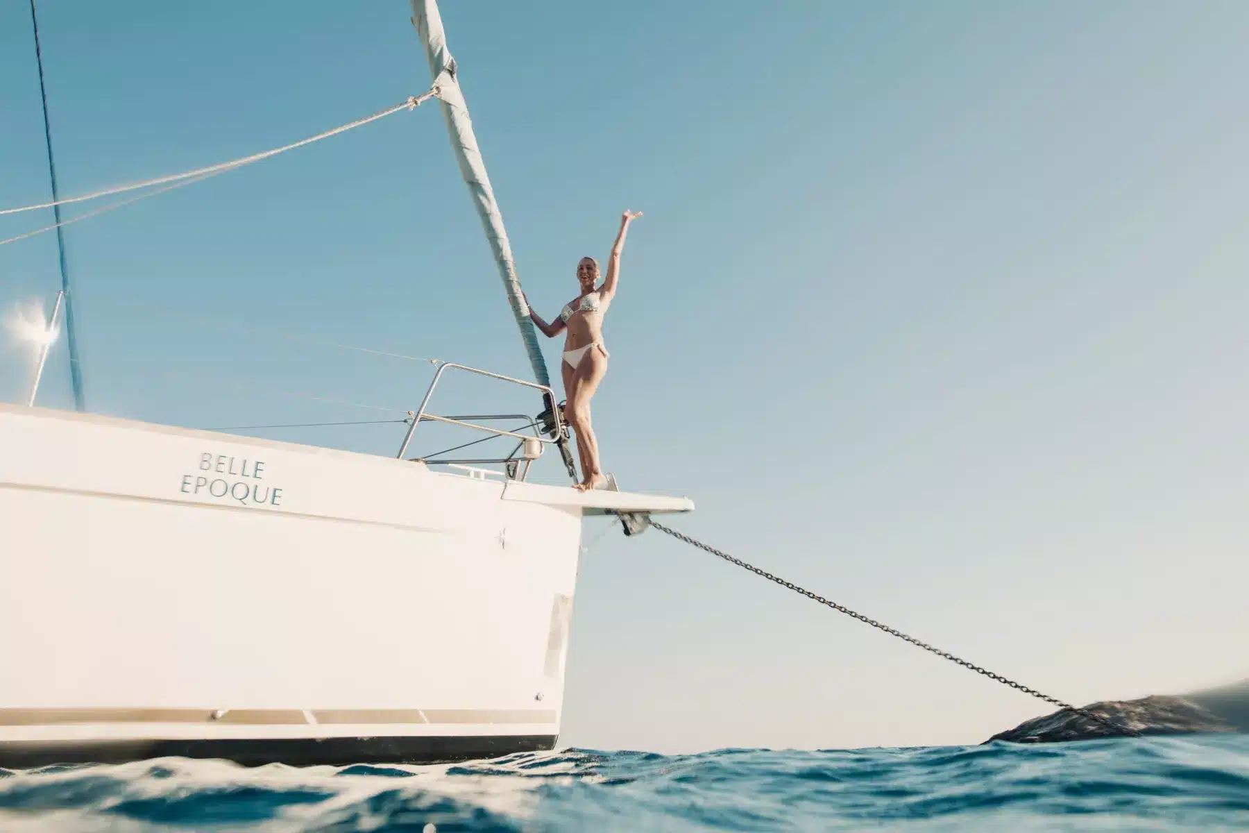 Eine Person streckt freudig die Arme in die Höhe, während sie am Bug eines Segelboots namens „Belle Epoque“ steht und während ihres Segelurlaubs durch ruhige, sonnenbeschienene Gewässer segelt.
