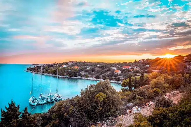 Ein ruhiger Strand bei Sonnenuntergang mit Segelyachten, die in einer ruhigen Bucht vor Anker liegen, umgeben von üppigem Grün unter einem farbenfrohen Himmel.