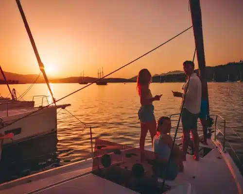 Eine Gruppe von Menschen genießt den Sonnenuntergang auf einer Segelyacht, mit ruhigem Wasser und anderen Booten im Hintergrund. Sie sind entspannt und unterhalten sich.
