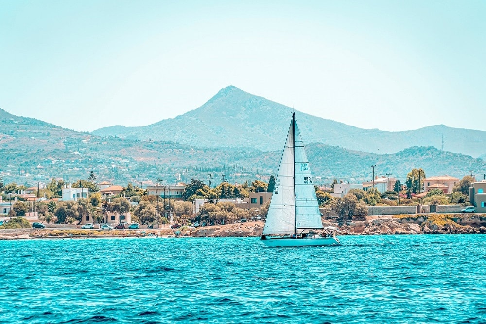 Ein Katamaran gleitet über türkisfarbenes Wasser in der Nähe einer Küstenstadt. Im Hintergrund ragt unter einem klaren blauen Himmel ein Berg auf.