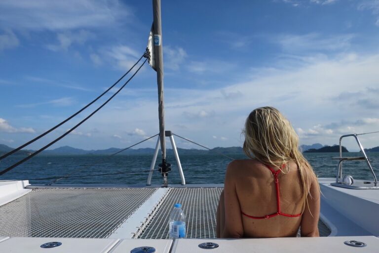 Eine Frau mit blondem Haar sitzt auf einer Segelyacht und blickt auf eine malerische Aussicht auf ferne Berge und einen wolkenverhangenen Himmel über einer großen Wasserfläche.