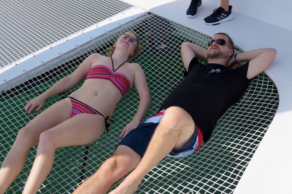 Zwei Personen sonnen sich auf dem Netz einer Segelyacht, eine trägt einen gestreiften Bikini und die andere ein schwarzes T-Shirt und Badeshorts und entspannt mit geschlossenen Augen.