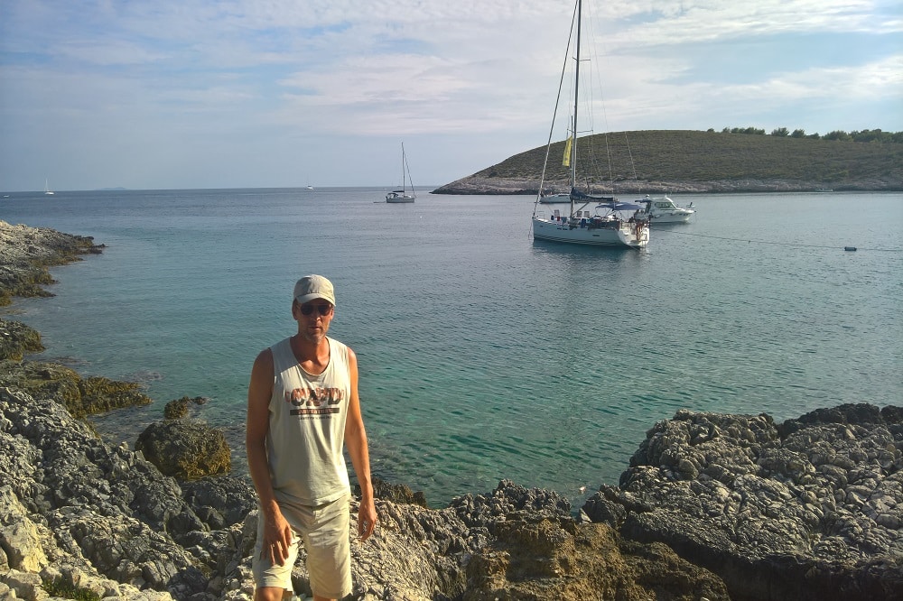 Ein Mann in Tanktop und Hut steht an einer felsigen Küste mit Segelbooten, die unter einem sonnigen Himmel im klaren blauen Wasser einer Bucht vor Anker liegen.