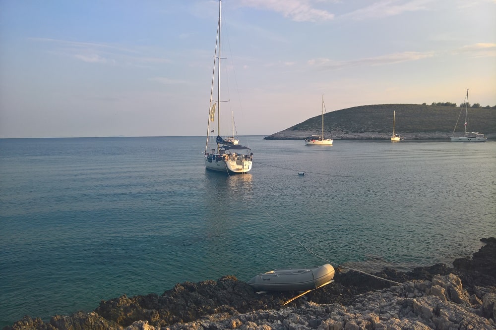 Eine ruhige Meereslandschaft mit einer Segelyacht, die in der Nähe einer felsigen Küste vor Anker liegt, mit einem Beiboot am Ufer und einer weiteren Jacht in der Ferne, unter einem sanften Abendhimmel.