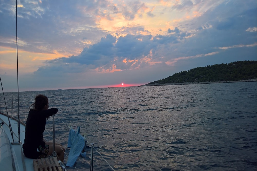 Eine Frau sitzt auf dem Deck einer Segelyacht und blickt auf den Sonnenuntergang über dem Meer, mit einer bewaldeten Küste in der Nähe.