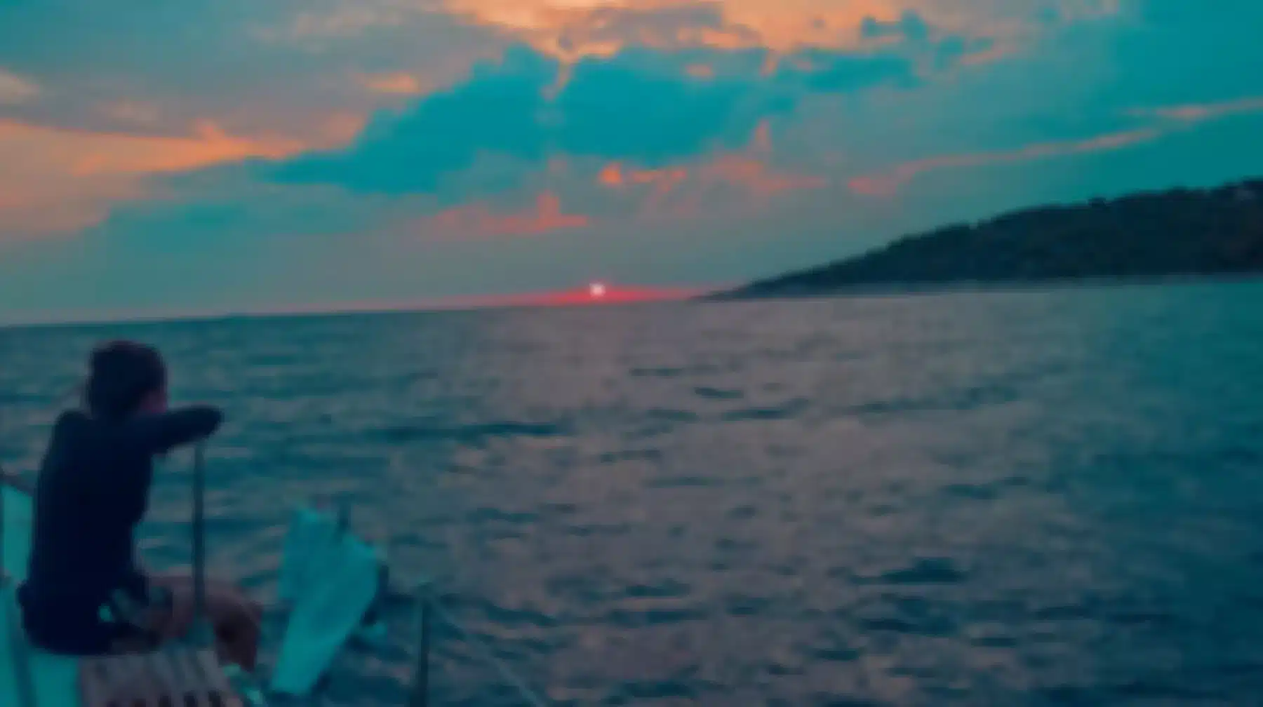 Eine Person sitzt während ihres Segelurlaubs auf einem Boot und blickt auf einen leuchtenden Sonnenuntergang über dem Meer, mit einem üppigen Hügel auf der rechten Seite, unter einem pastellblauen und rosa Himmel.