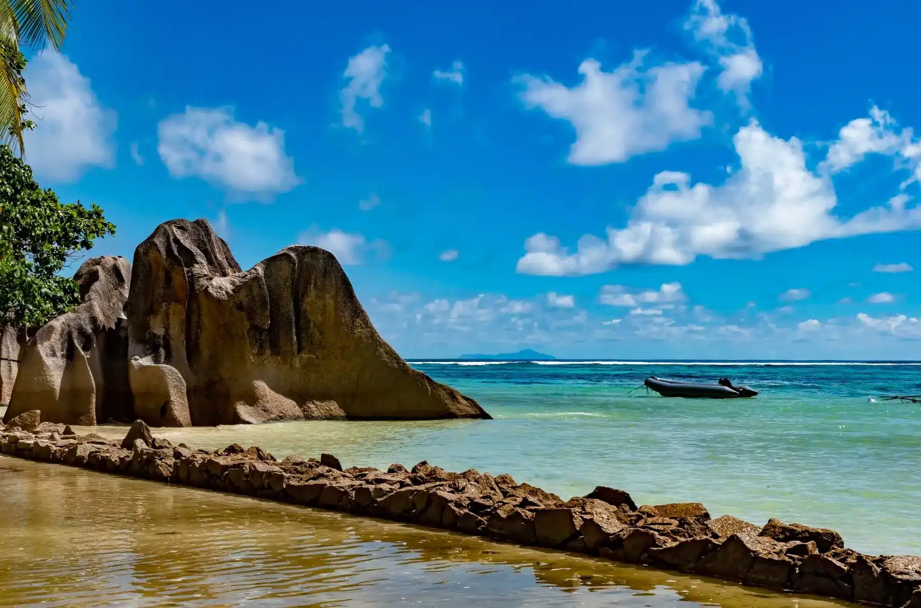 Eine malerische Aussicht auf große Granitfelsen an einem tropischen Strand mit kristallklarem, türkisfarbenem Wasser unter einem blauen Himmel und einer kleinen Segelyacht, die in der Ferne schwimmt.