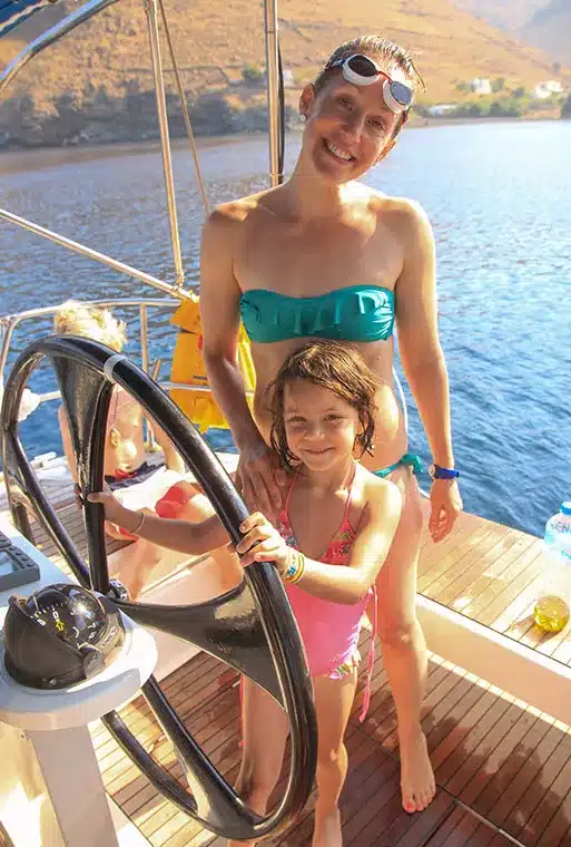 Eine Frau in einem blaugrünen Bandeau-Bikinioberteil und ein Mädchen in einem rosa einteiligen Badeanzug lächeln neben einem Schiffssteuerrad auf einer Segelyacht an einem sonnigen Tag mit Hügeln im